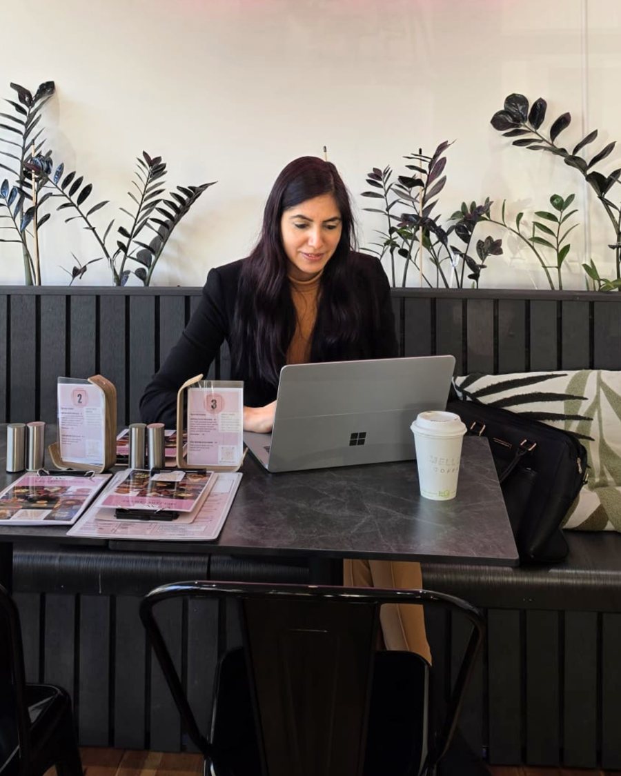 Kanika Vasudeva working on her laptop at a cafe table, with a coffee beside her.